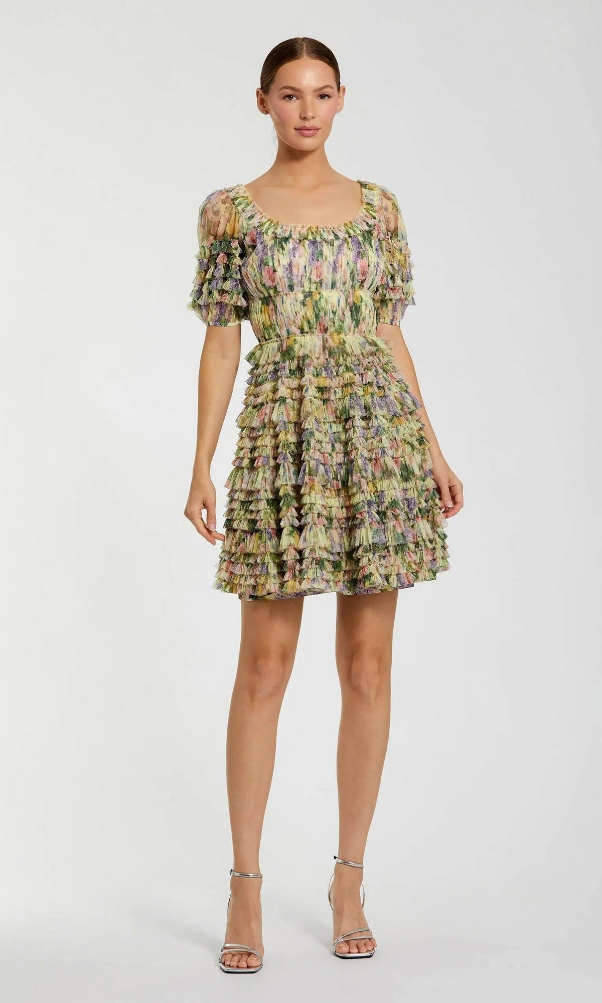A woman stands in a studio, facing forward against a plain light background, wearing the Mac Duggal 8327 Short Party Dress in Yellow Multi—an eye-catching mini dress adorned with multicolored ruffles and short sleeves—paired with silver high-heeled sandals.